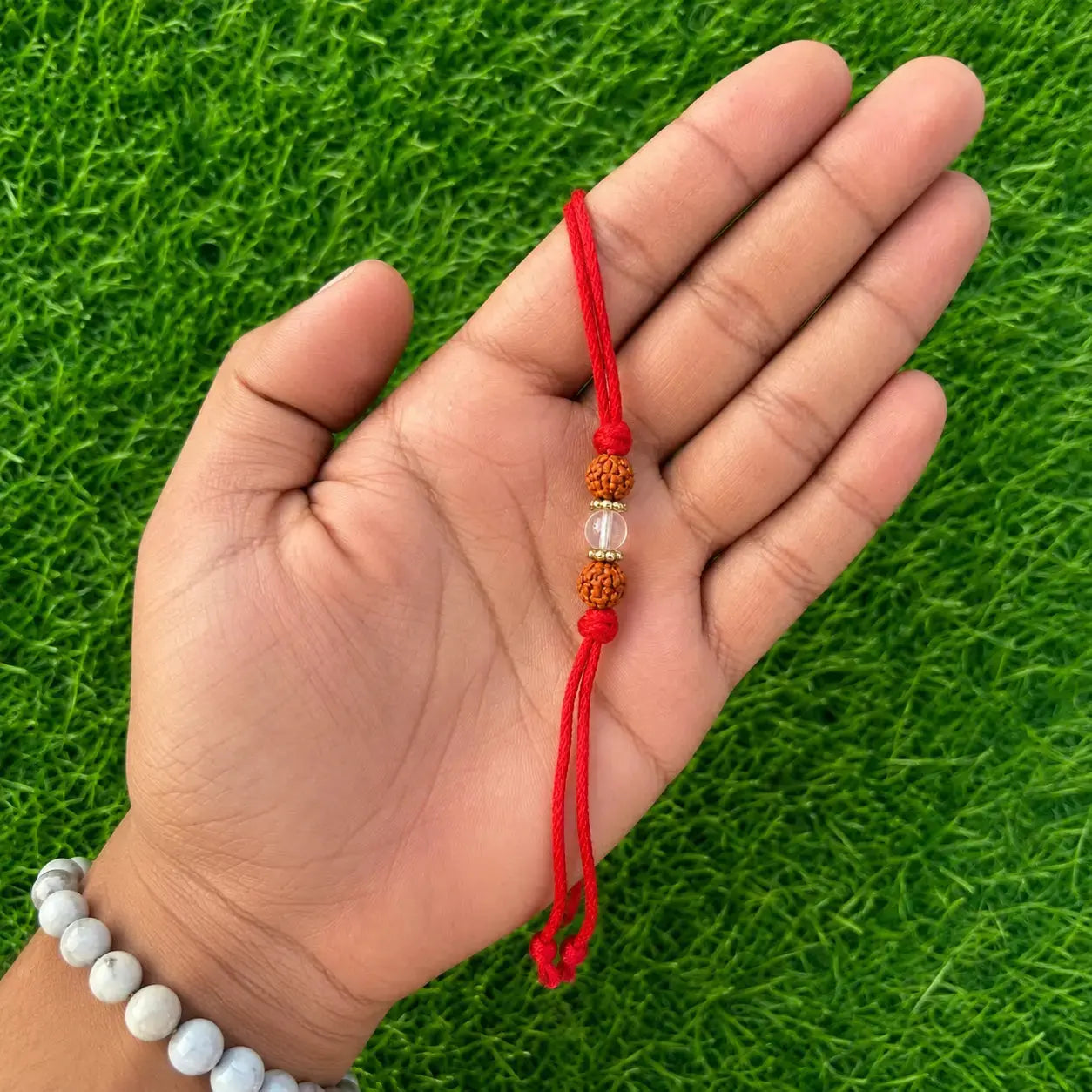 Hand holding a red string bracelet with beads against a green grass background